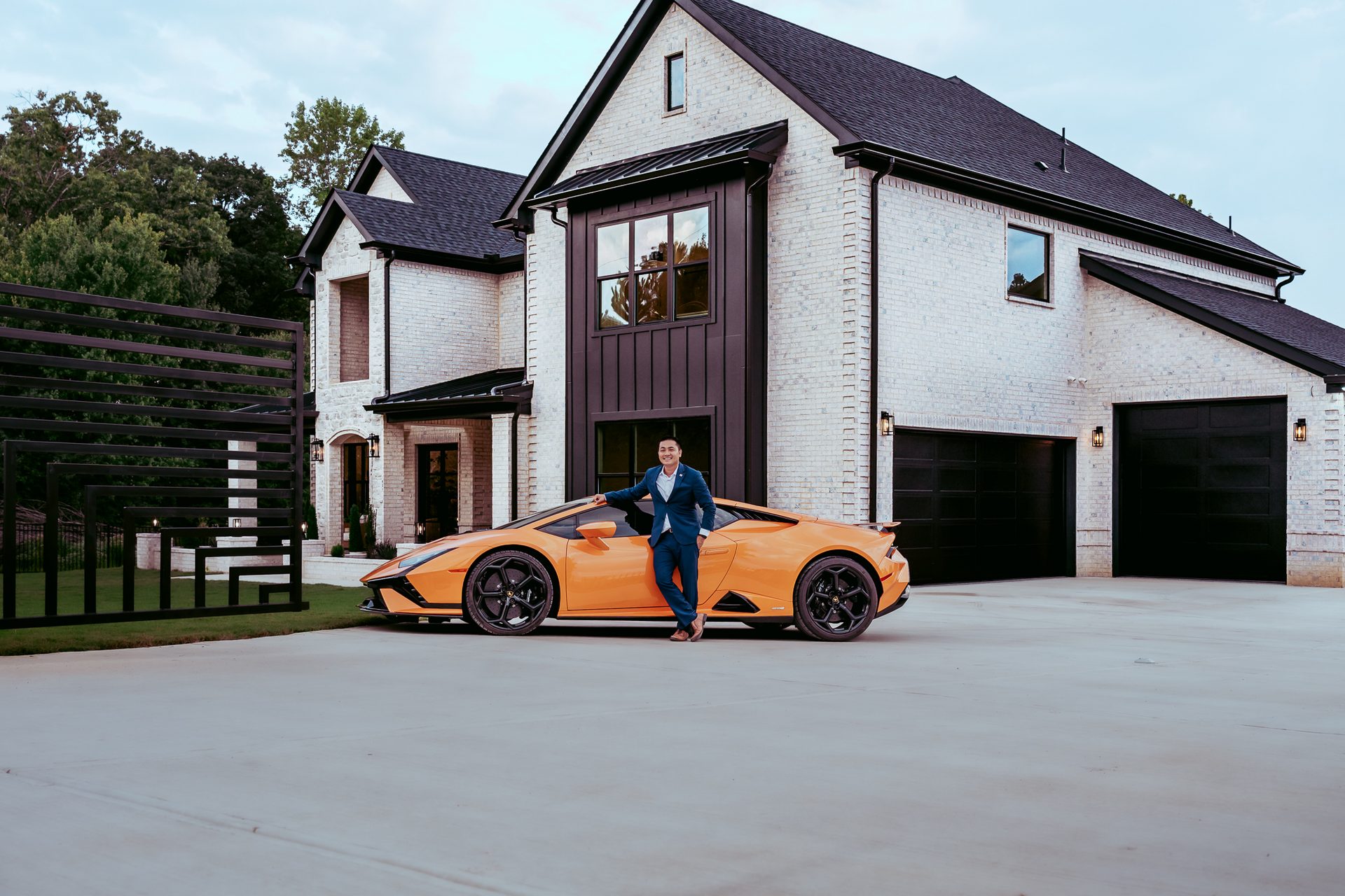 Túy “Dewey” Luong in a navy suit beside an orange sports car at a luxury home during his brand shoot