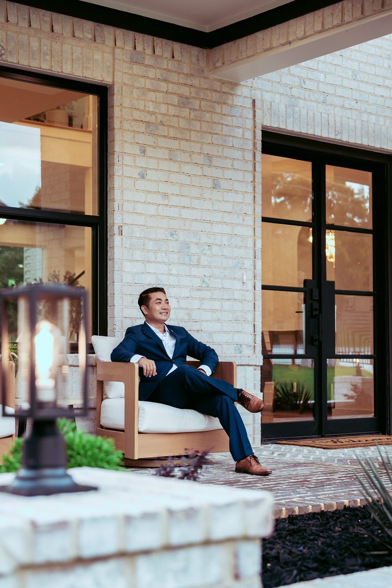 Dewey Luong in a navy suit sitting relaxed in a wooden chair on the porch of a luxury home during his 2026 brand shoot